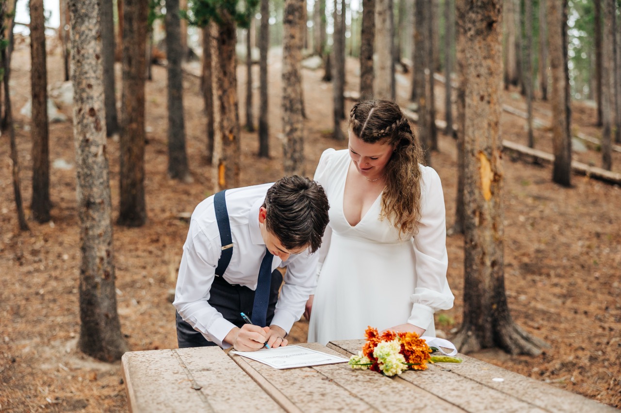 Bride watches as groom signs their marriage license during their decision to elope in Boulder’s pine forest.
