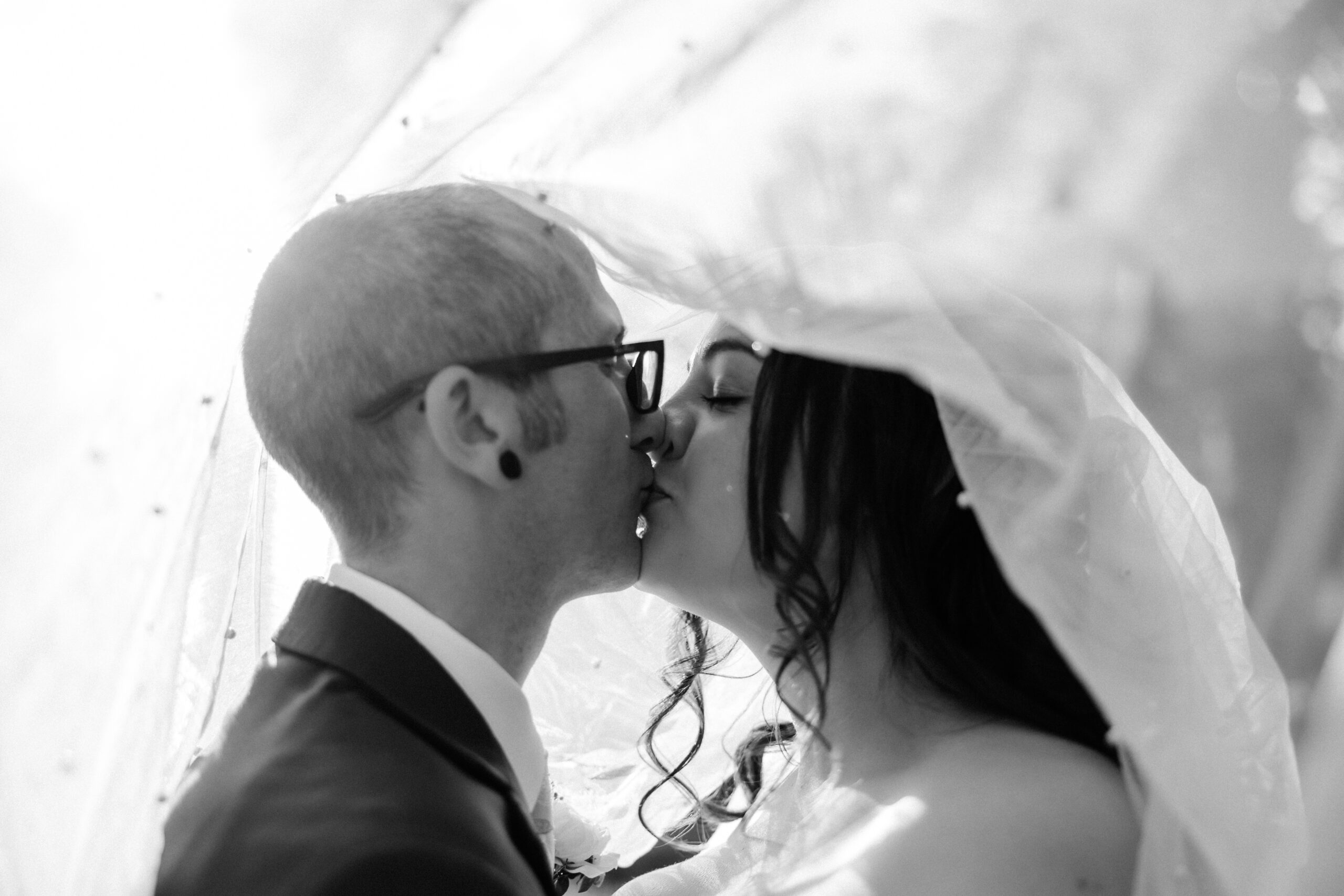 Couple shares a soft, romantic kiss beneath the bride’s veil, bathed in gentle light and emotion.