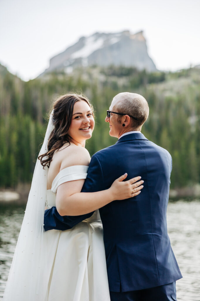 Bride and partner smile and embrace among boulders and budding trees, with a lake and mountain landscape behind them.