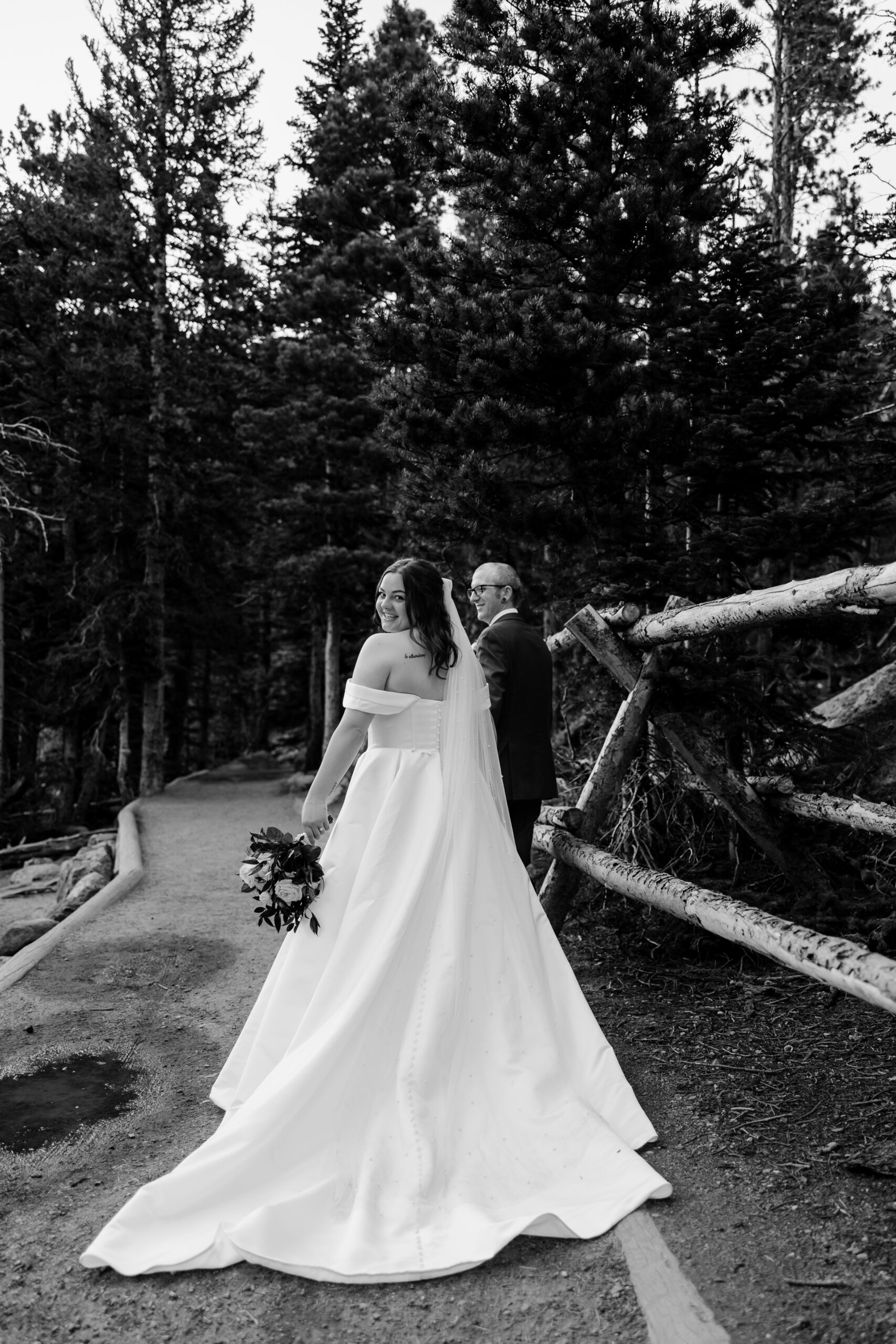 Bride glances back with a joyful smile while walking a forest trail beside her partner, holding a bouquet of roses.