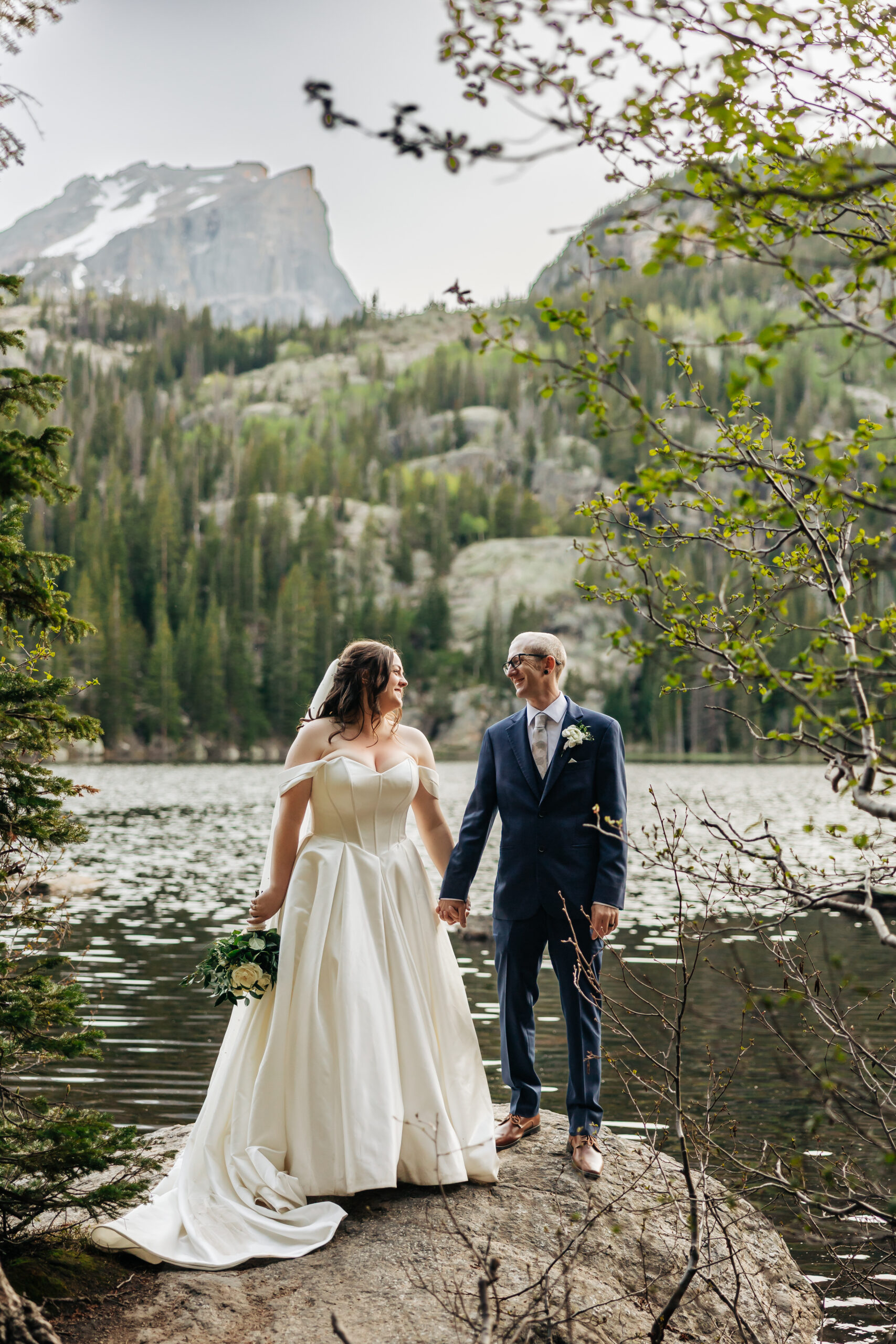 Bride and groom hold hands on a rocky lakeside outcrop beneath a towering mountain, beautifully framed by a Denver elopement photographer.