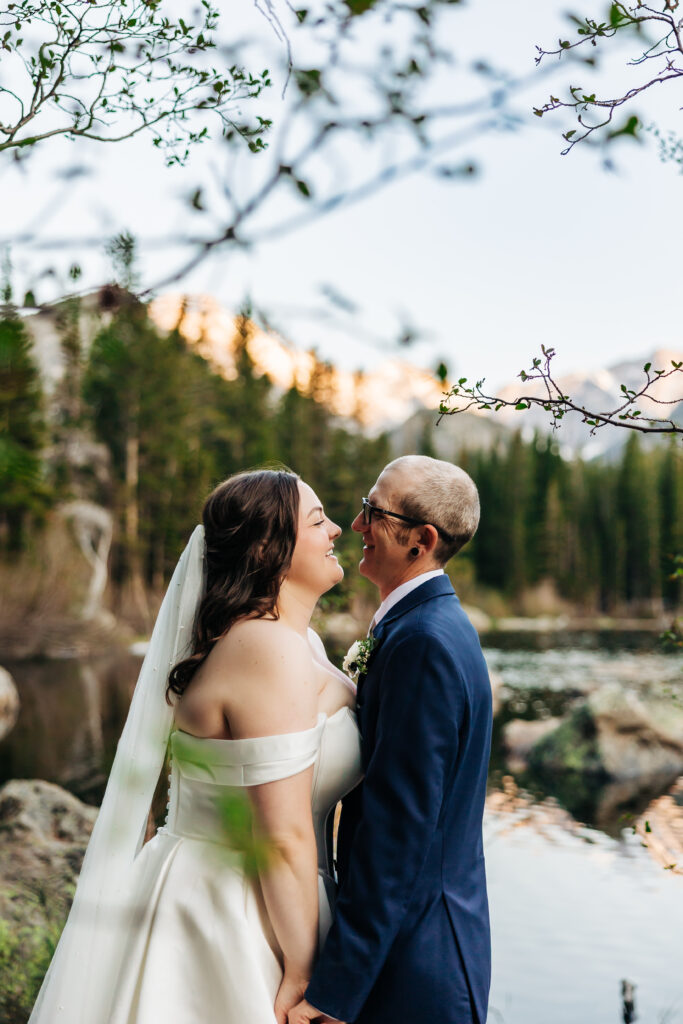 Couple stands close, smiling and gazing into each other’s eyes on the shore of a quiet lake at sunset.