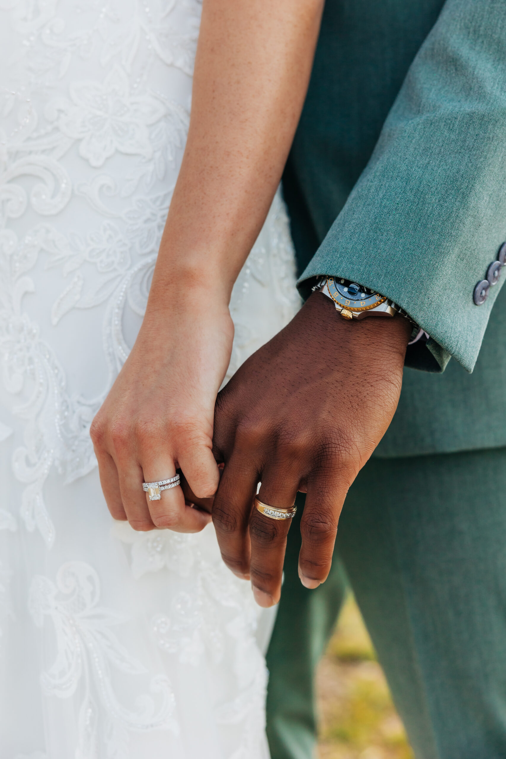Close-up of bride and groom holding hands, their wedding rings shining against lace and a sage green suit during their Colorado mountain wedding.
