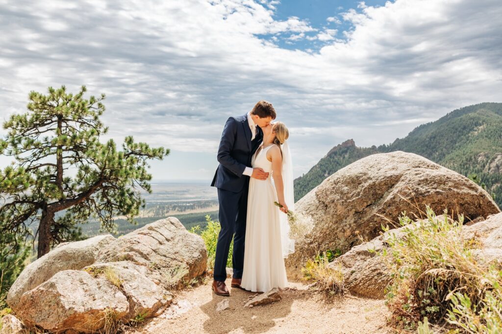 Newlyweds share a kiss in front of rugged boulders and sweeping views, soaking in the beauty of their wedding day hike adventure.