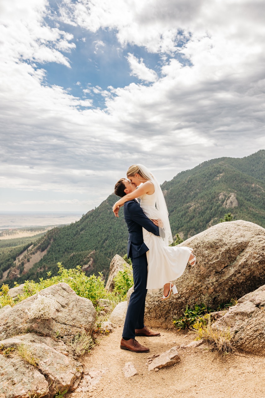 Groom lifts his bride off the ground for a joyful kiss, surrounded by dramatic mountain peaks—an unforgettable moment from their wedding day hike.