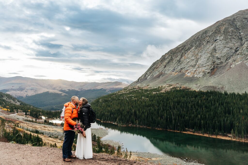 couple hiking while getting married. there's a lake and mountains in the background. the couple is kissing
