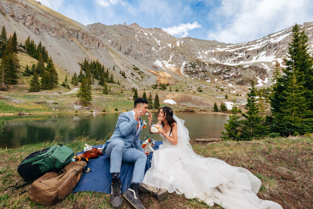A bride and groom feeding each other cake while having a picnic with a lake and mountains in the back for their adventure elopement
