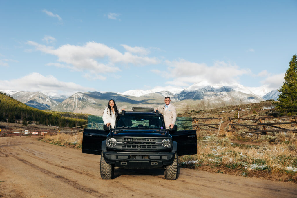 A couple standing in their car with mountains in the background for their adventure elopement