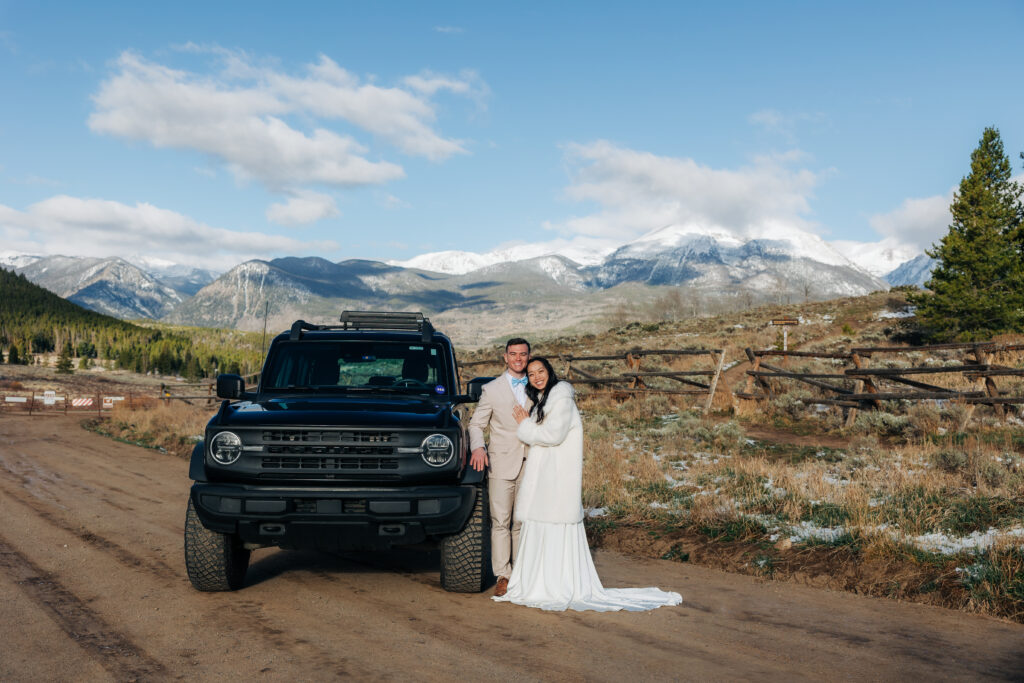 A couple standing next to a Ford Bronco for their off-roading elopement. There are mountains in the background with snow. 