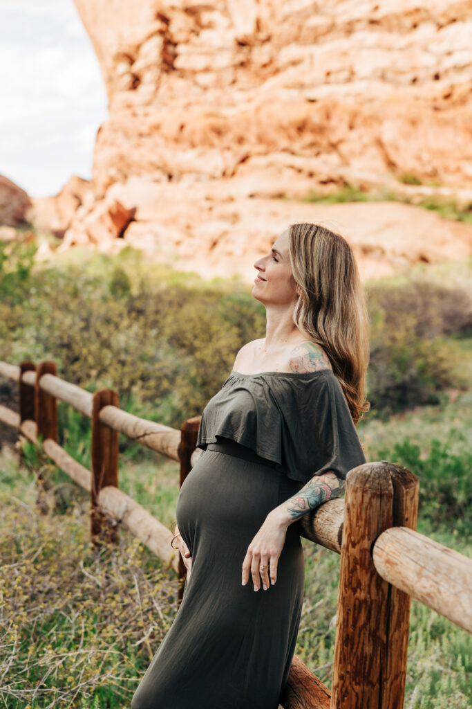 Denver Maternity Photographer captures woman leaning against the fence wearing olive green maternity dress