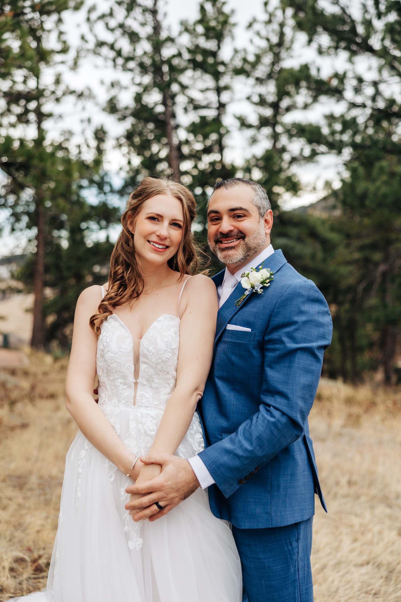 miling bride and groom hold hands during their outdoor Colorado wedding, surrounded by tall pine trees and rustic forest scenery.