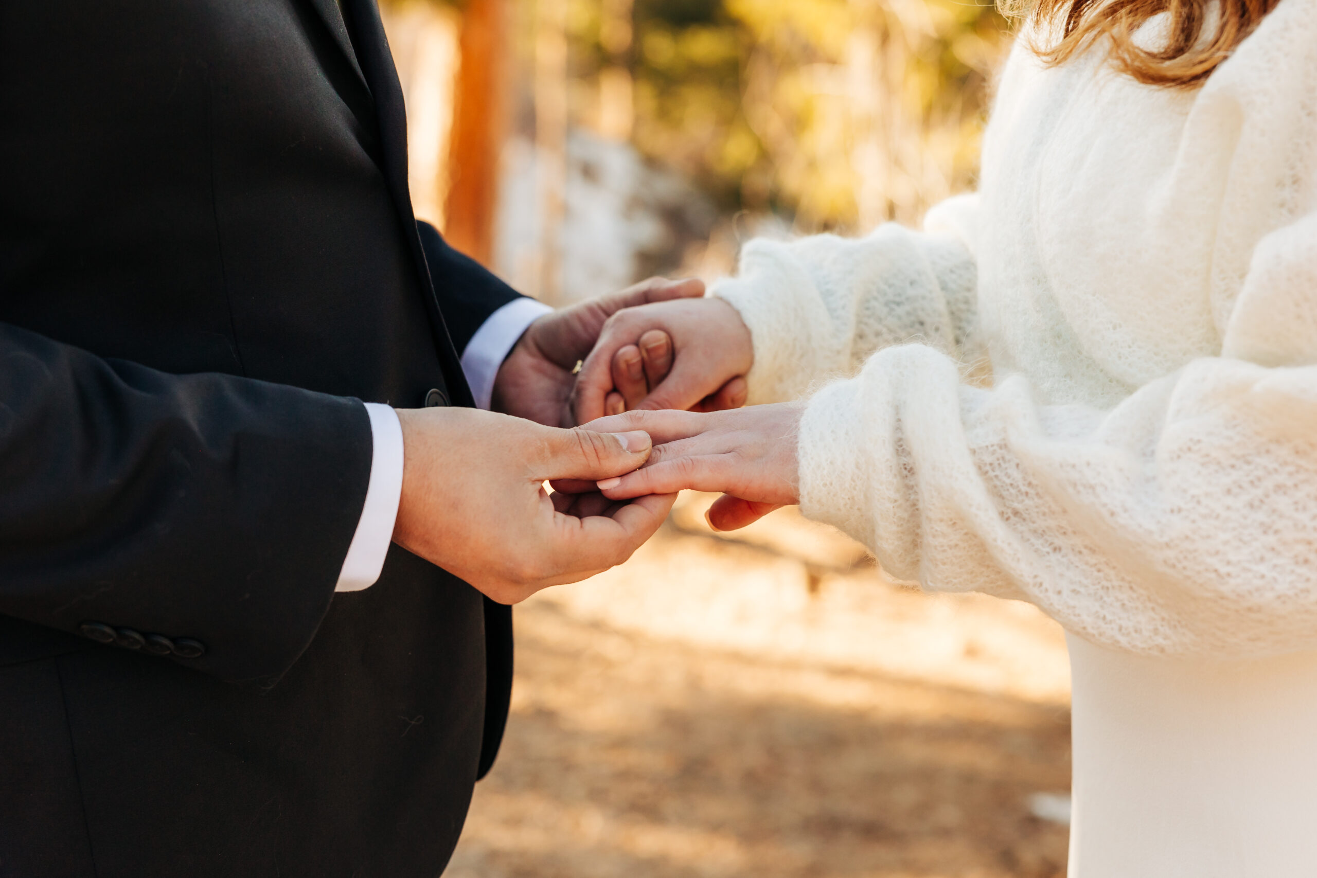 Groom places a wedding ring on his bride’s finger, their hands framed by warm afternoon light and soft knit textures.