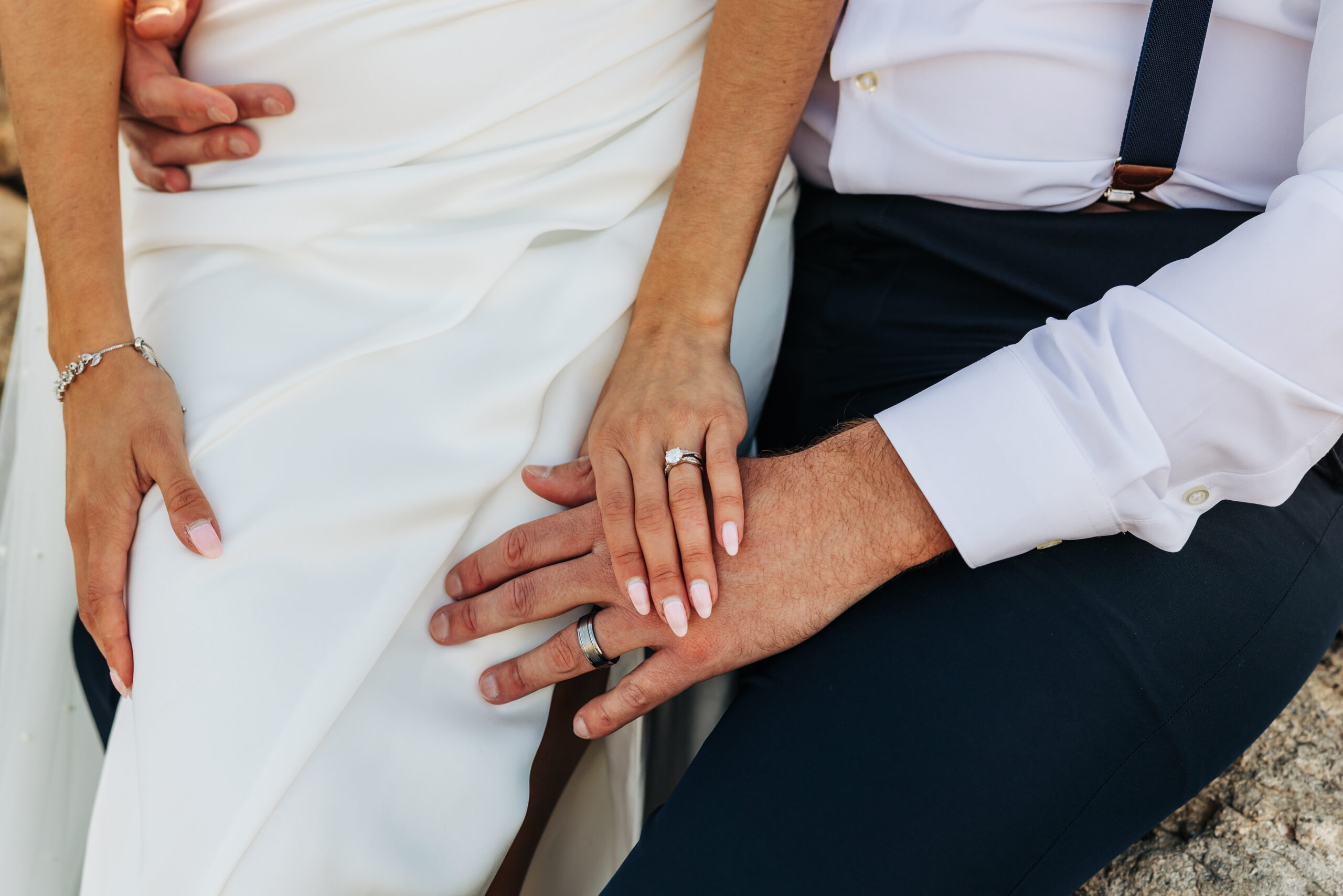 Close-up of couple’s hands and wedding rings resting together on the bride’s gown.