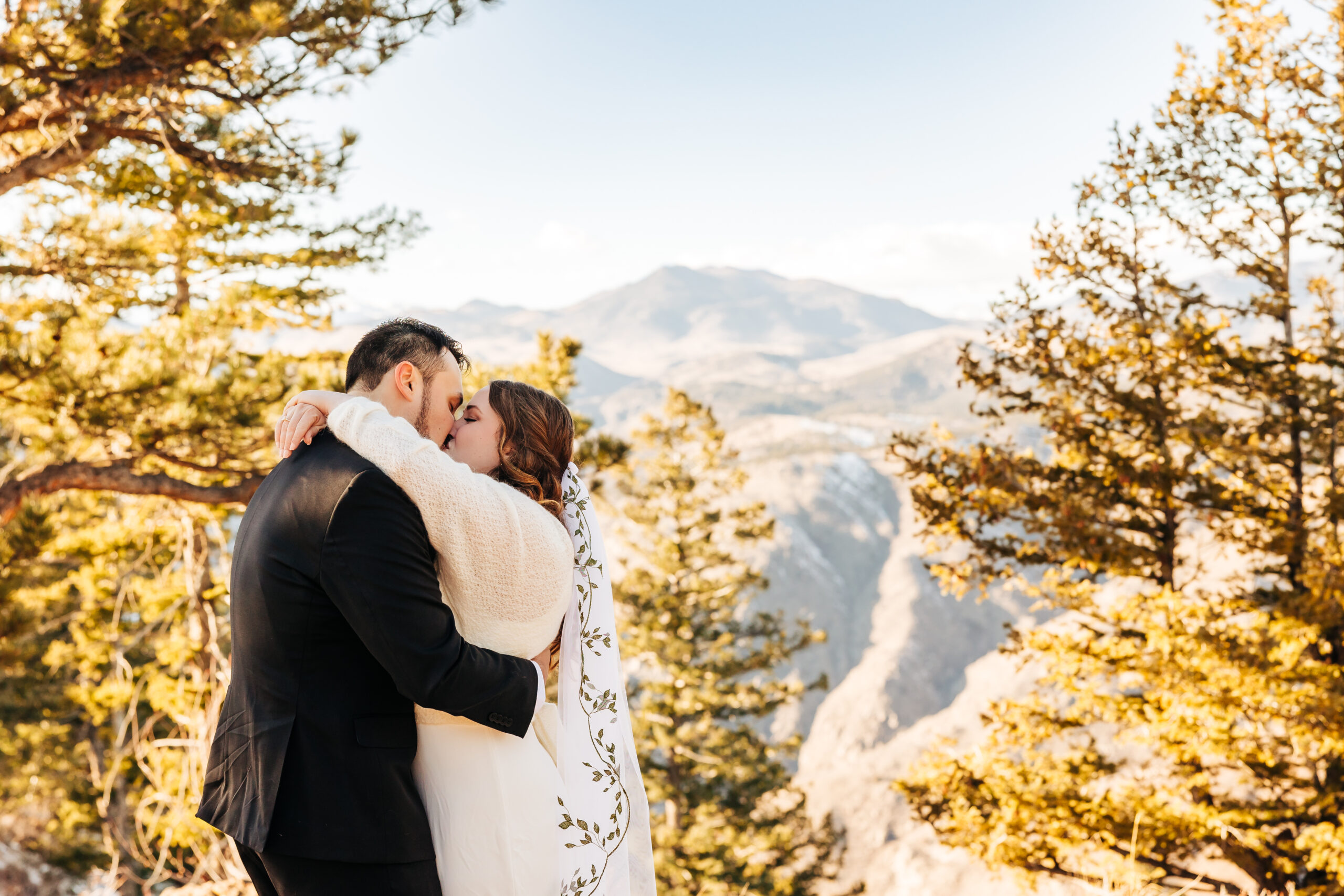 Bride and groom stand smiling hand-in-hand with their pup in the forest, surrounded by sun-dappled trees at their Boulder wedding.