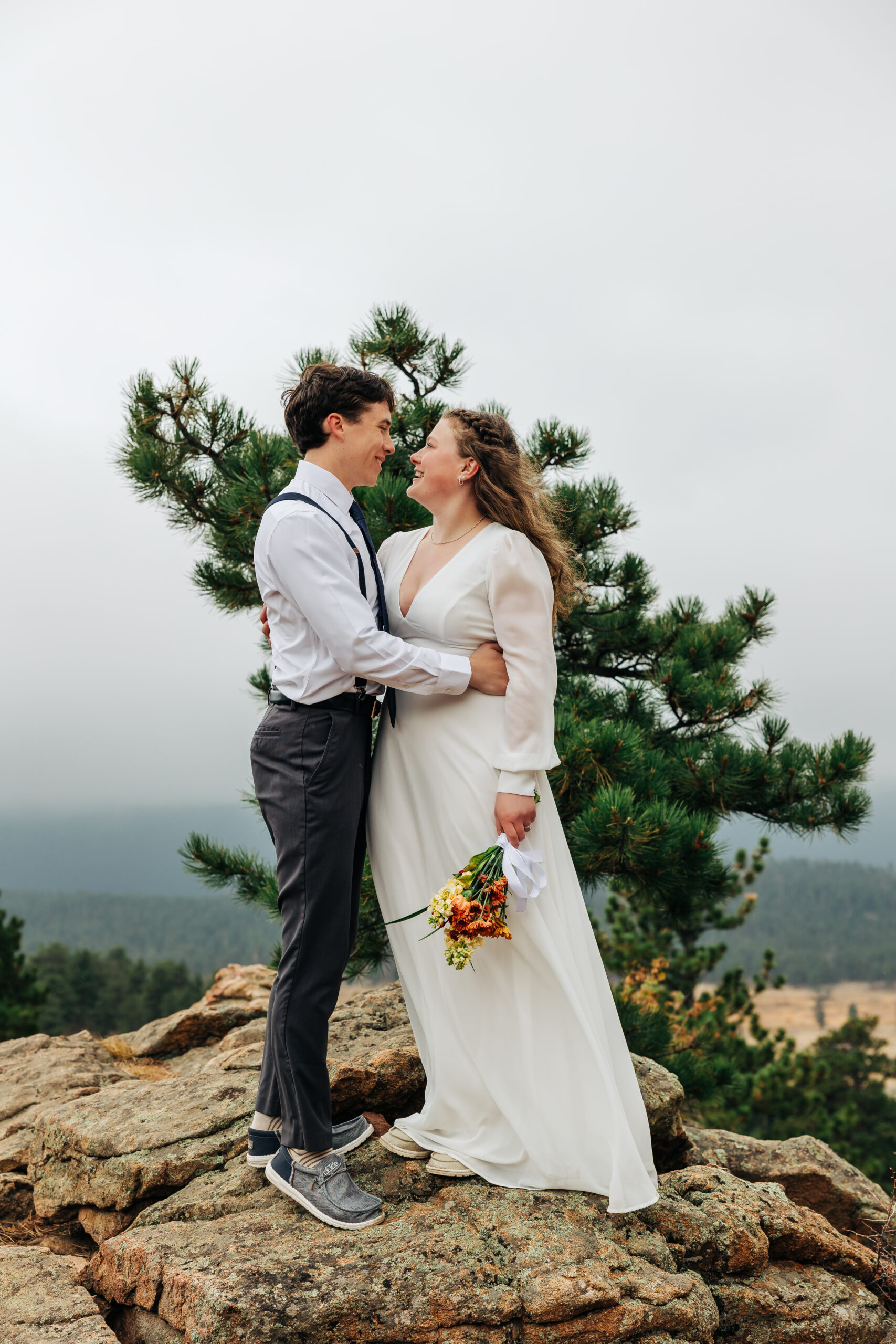 Couple stands on a rock ledge surrounded by evergreens, celebrating their Colorado elopement.