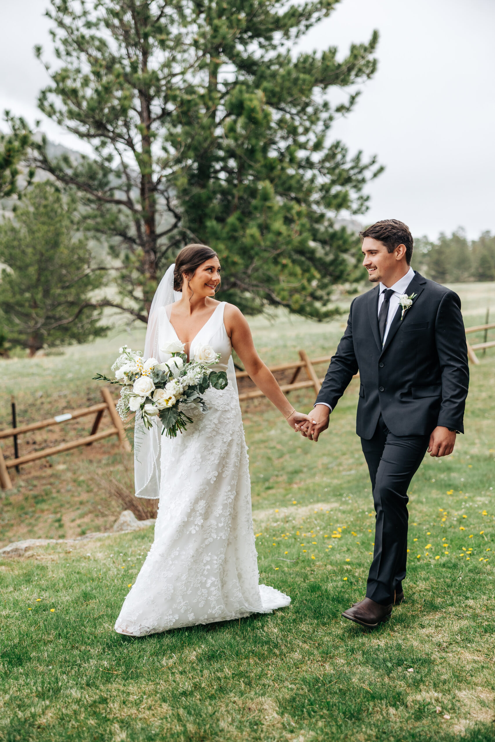 Bride and groom walk hand-in-hand across a grassy field, smiling at each other with pine trees in the background.