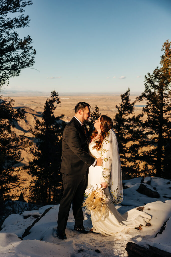 Couple shares a romantic kiss framed by pine trees and snow patches with distant mountain peaks behind them.
