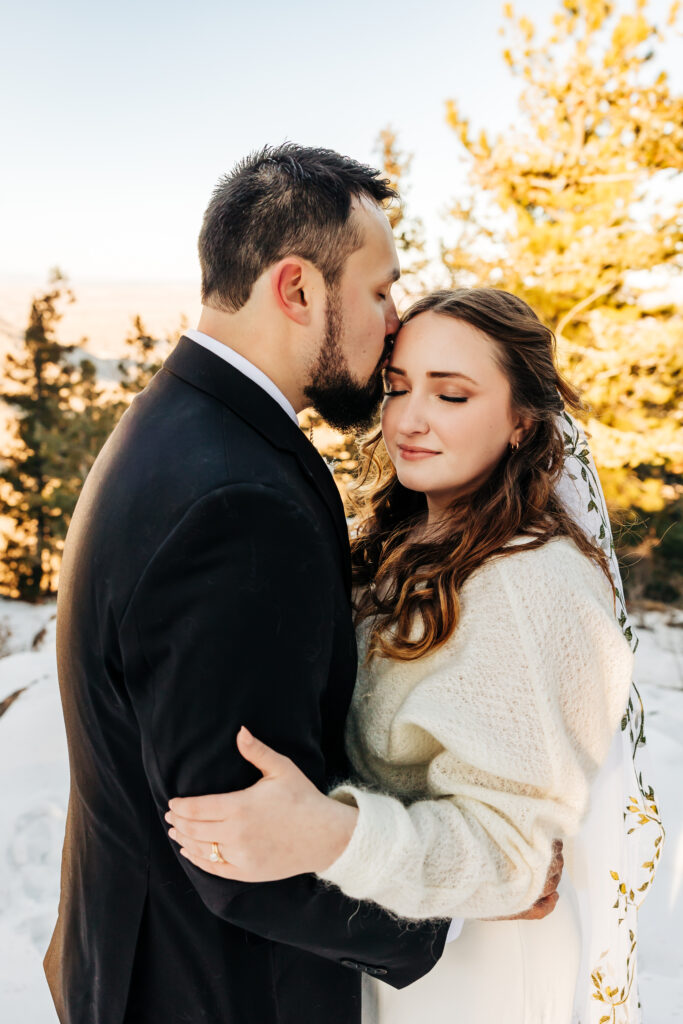 Groom kisses bride’s forehead as she closes her eyes peacefully, surrounded by soft snow and golden evergreens.