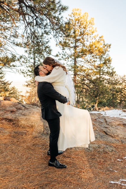 Groom lifts bride off the ground in a joyful embrace during their winter Boulder wedding surrounded by pine trees and golden light.