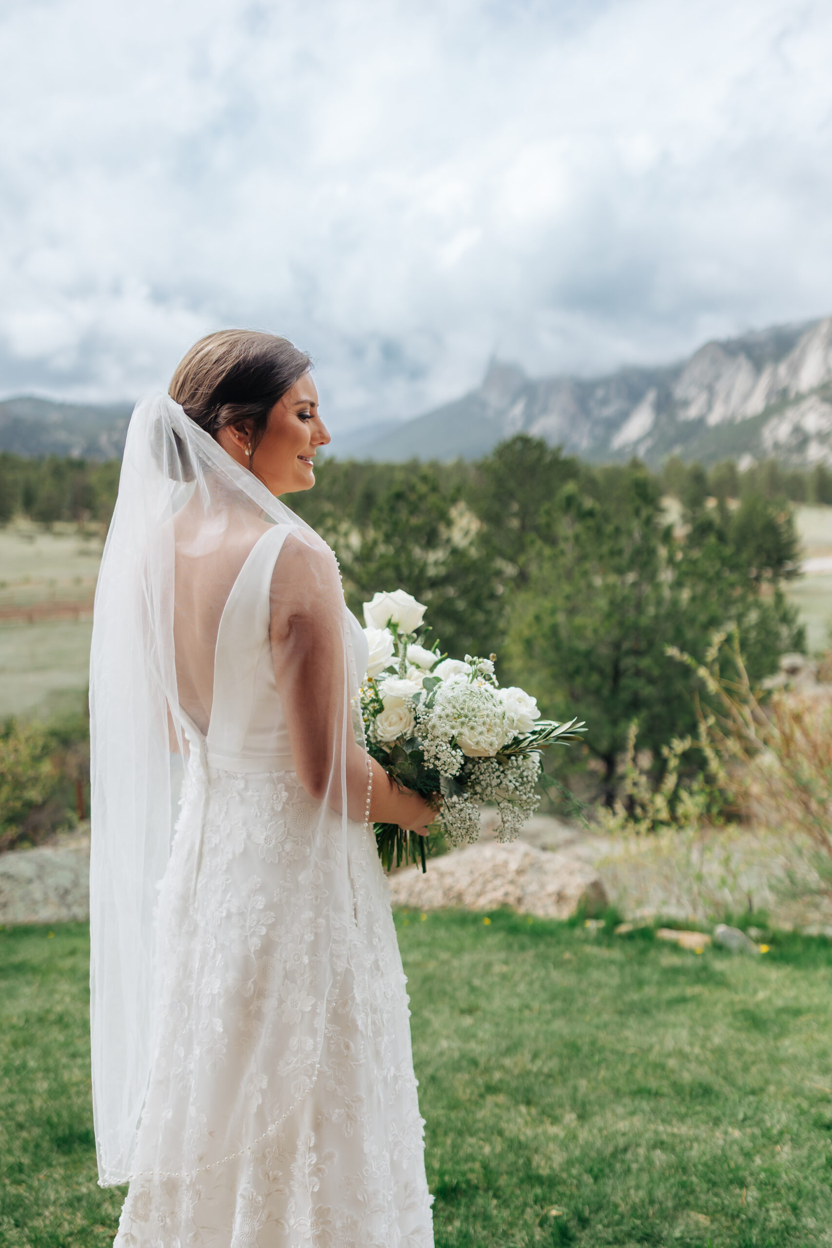 Bride in a floral lace gown holding a bouquet of white flowers, gazes out over the scenic mountain backdrop during her outdoor Colorado wedding.