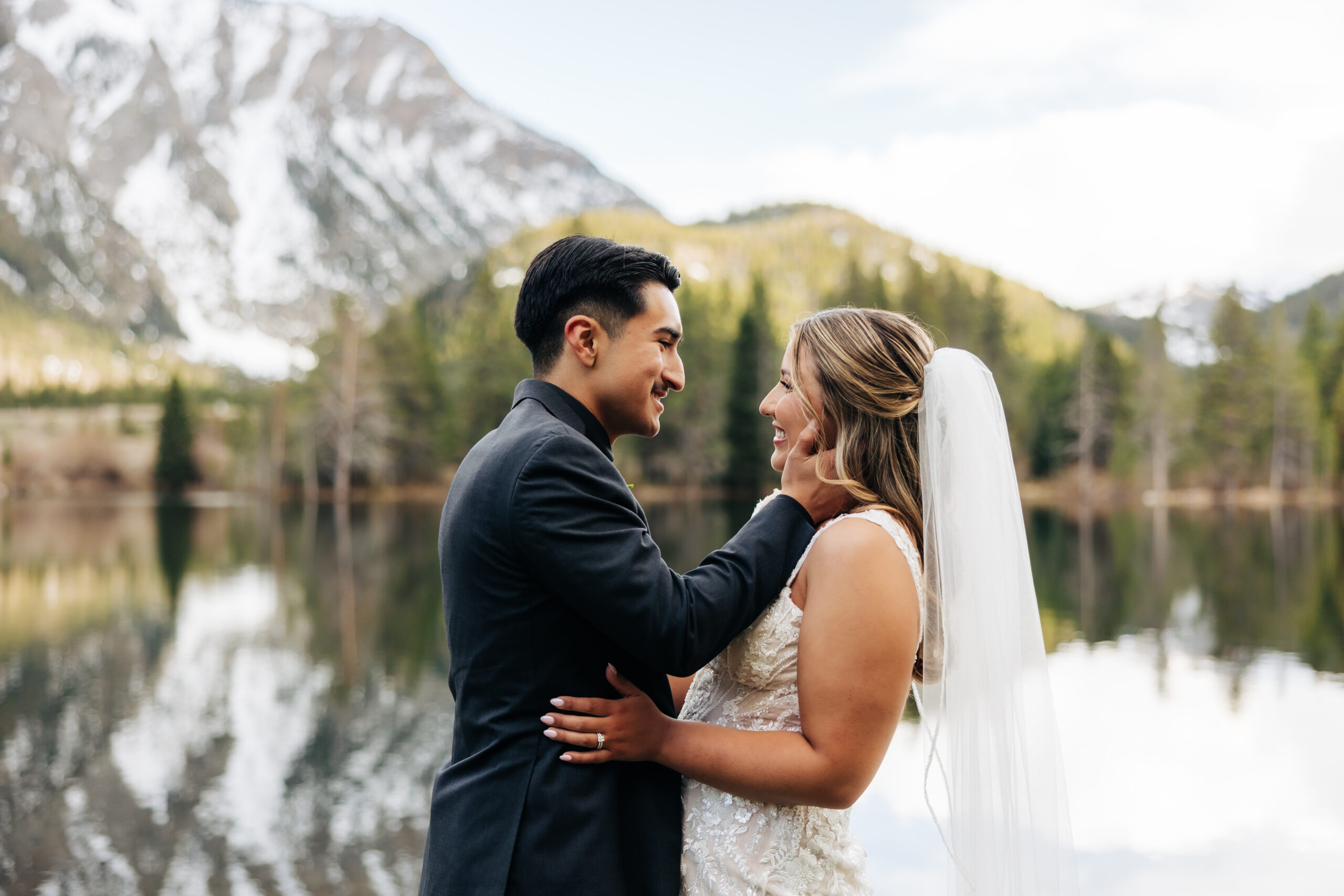 Bride and groom embrace by a peaceful alpine lake with snowy mountains in the background during their outdoor Colorado wedding.