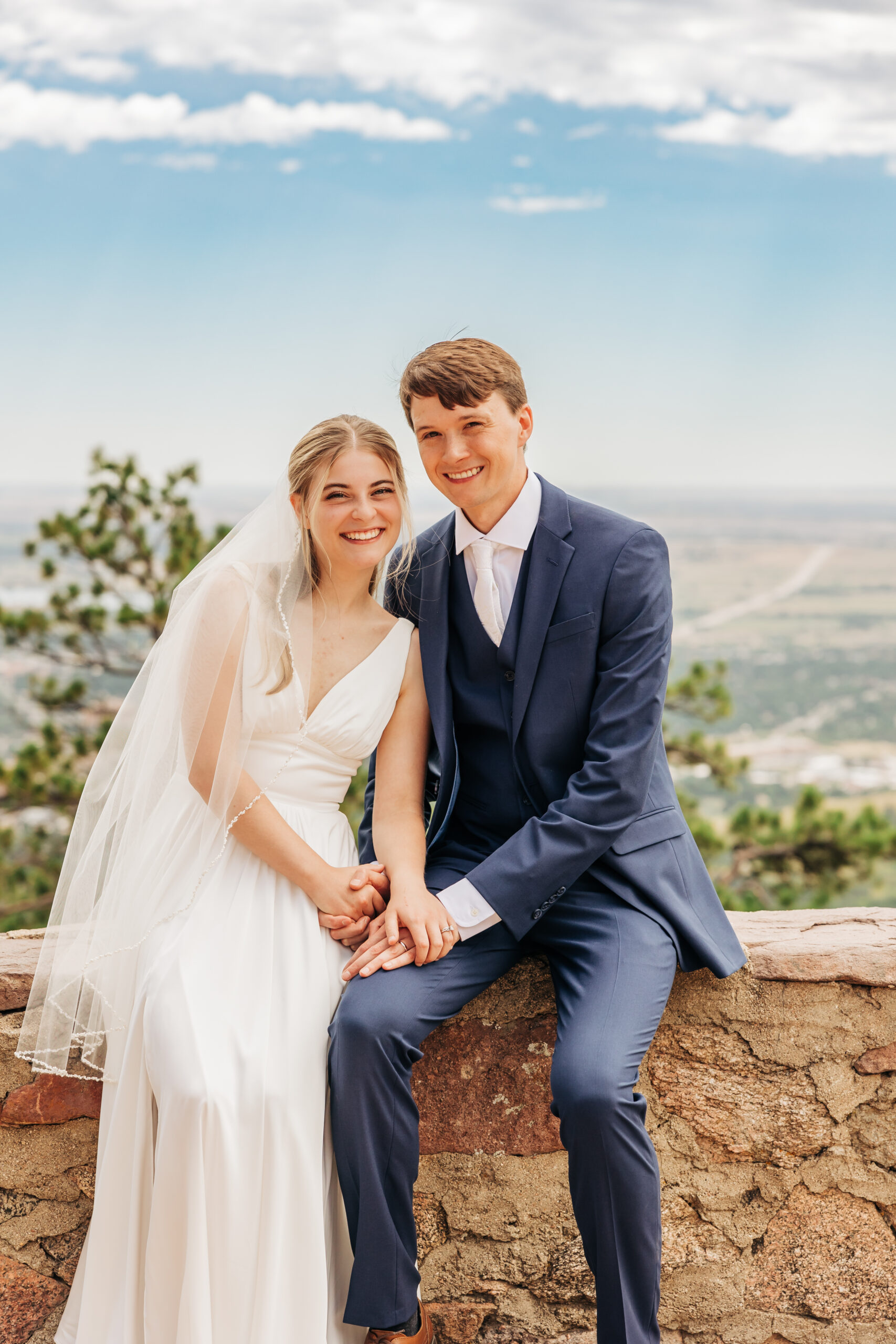Bride and groom sit hand in hand on a stone ledge, smiling with the plains stretching out behind them—a joyful moment from their Colorado wedding day hike.