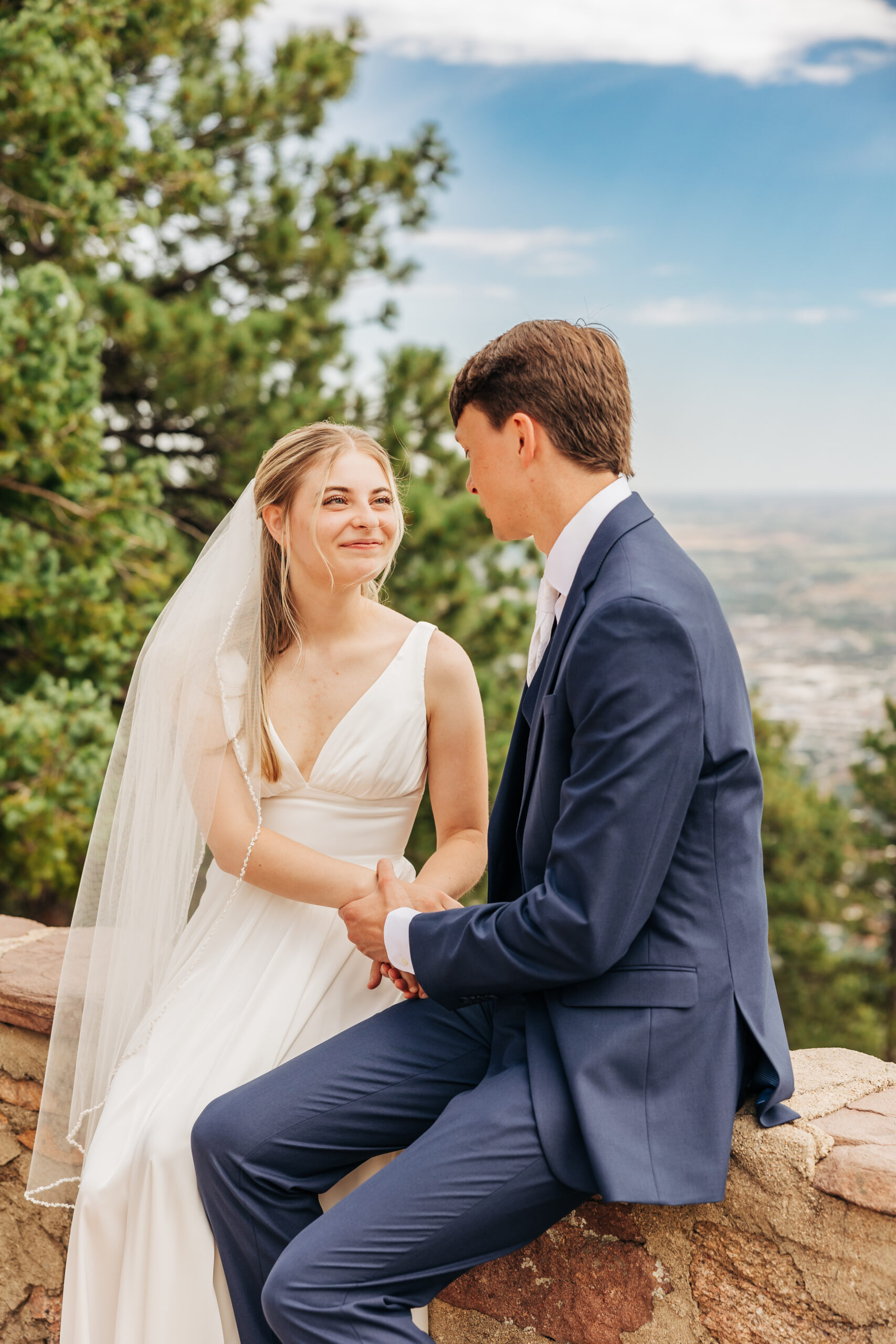 Holding hands on a stone wall, the bride and groom smile at one another with forest and sky as their backdrop—true connection on their wedding day hike.