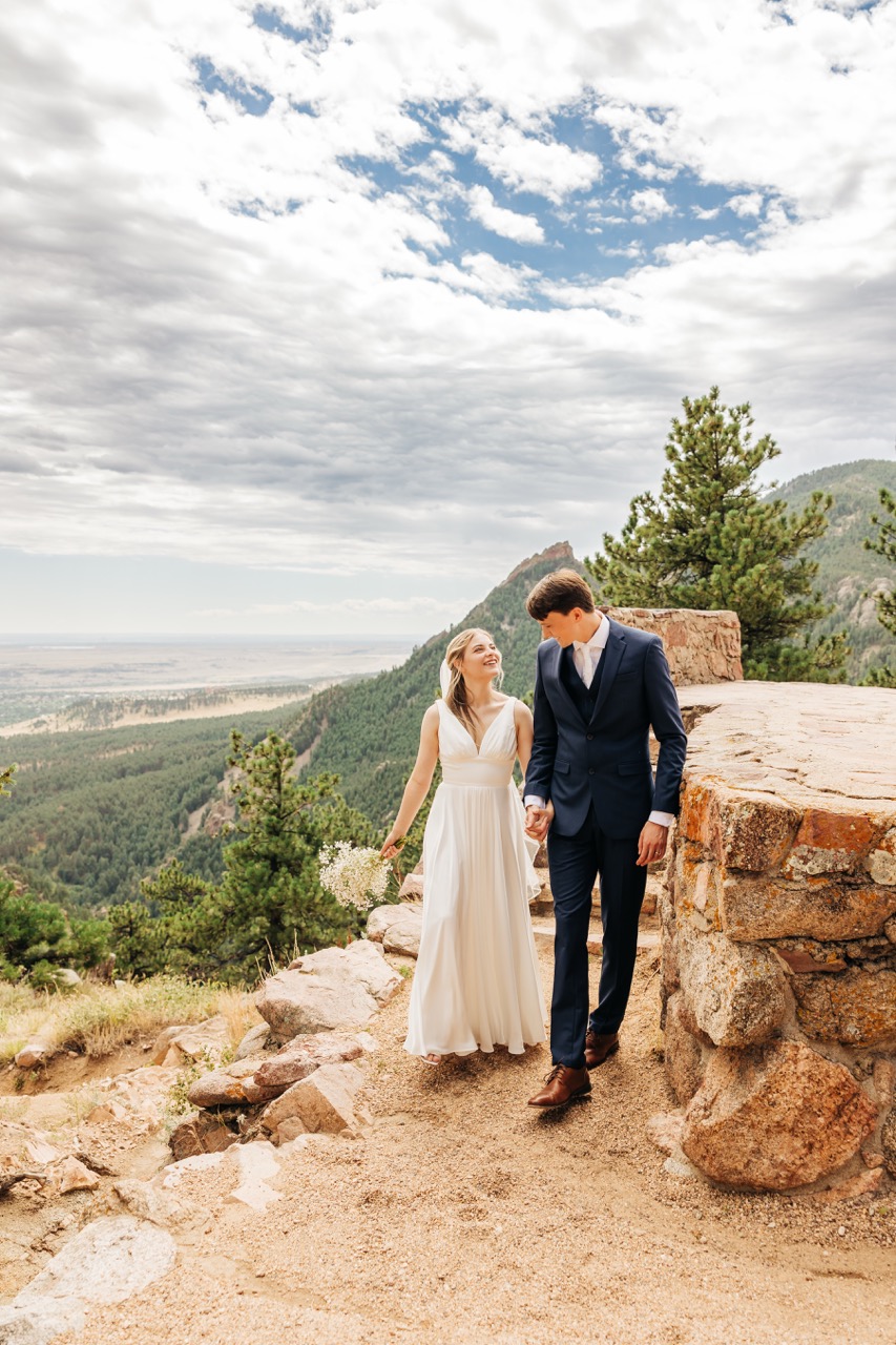 Bride and groom walk hand in hand along a stone path with sweeping mountain views behind them, sharing a joyful moment during their Colorado wedding day hike.