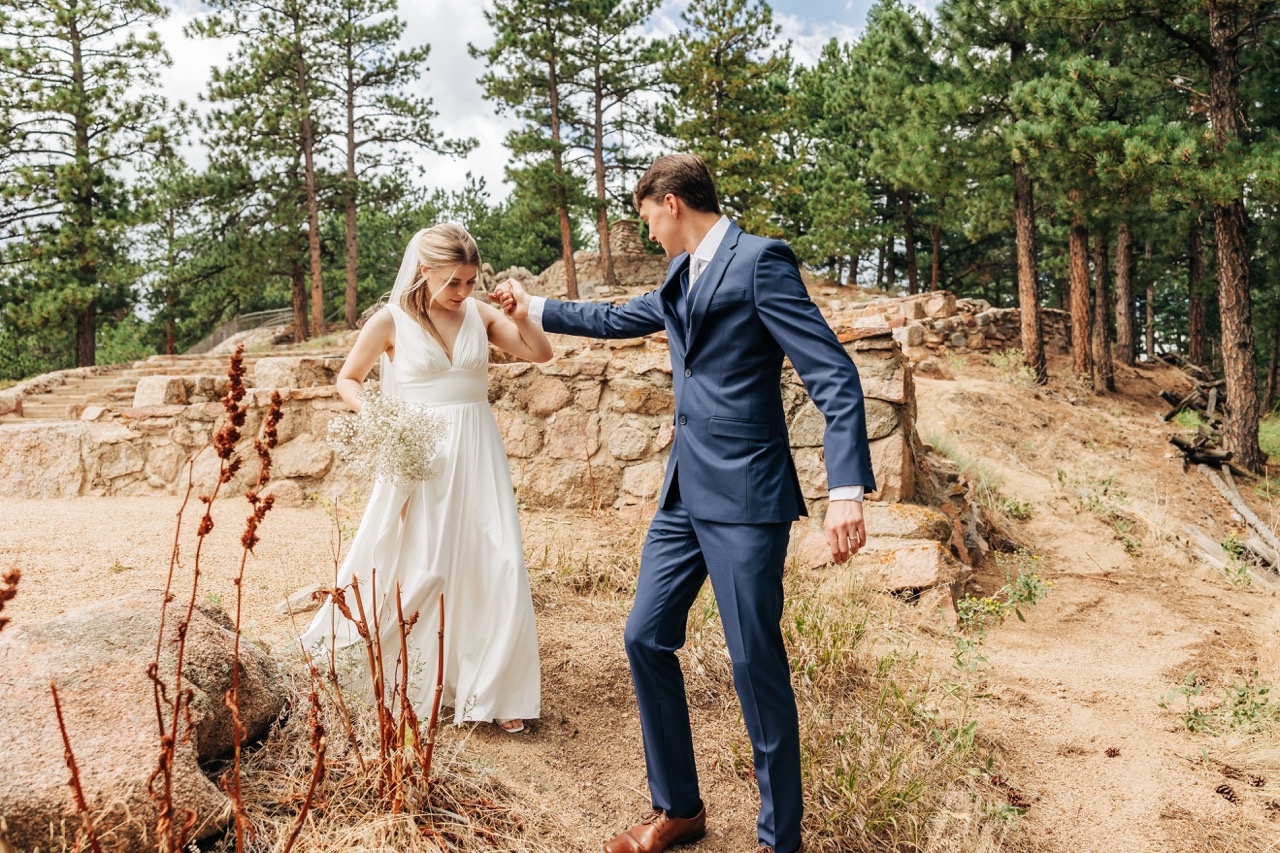 Groom offers a hand to help his bride along a dirt path lined with wildflowers and stone ruins—romance and teamwork on their wedding day hike.