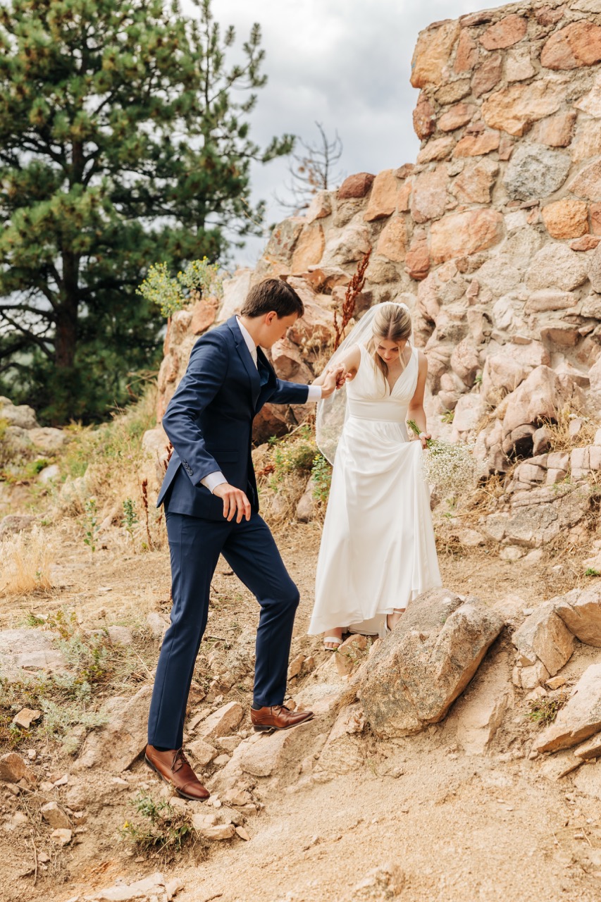 Bride carefully steps down a rocky trail with her groom guiding her hand, capturing a candid, tender moment from their wedding day hike in Colorado.