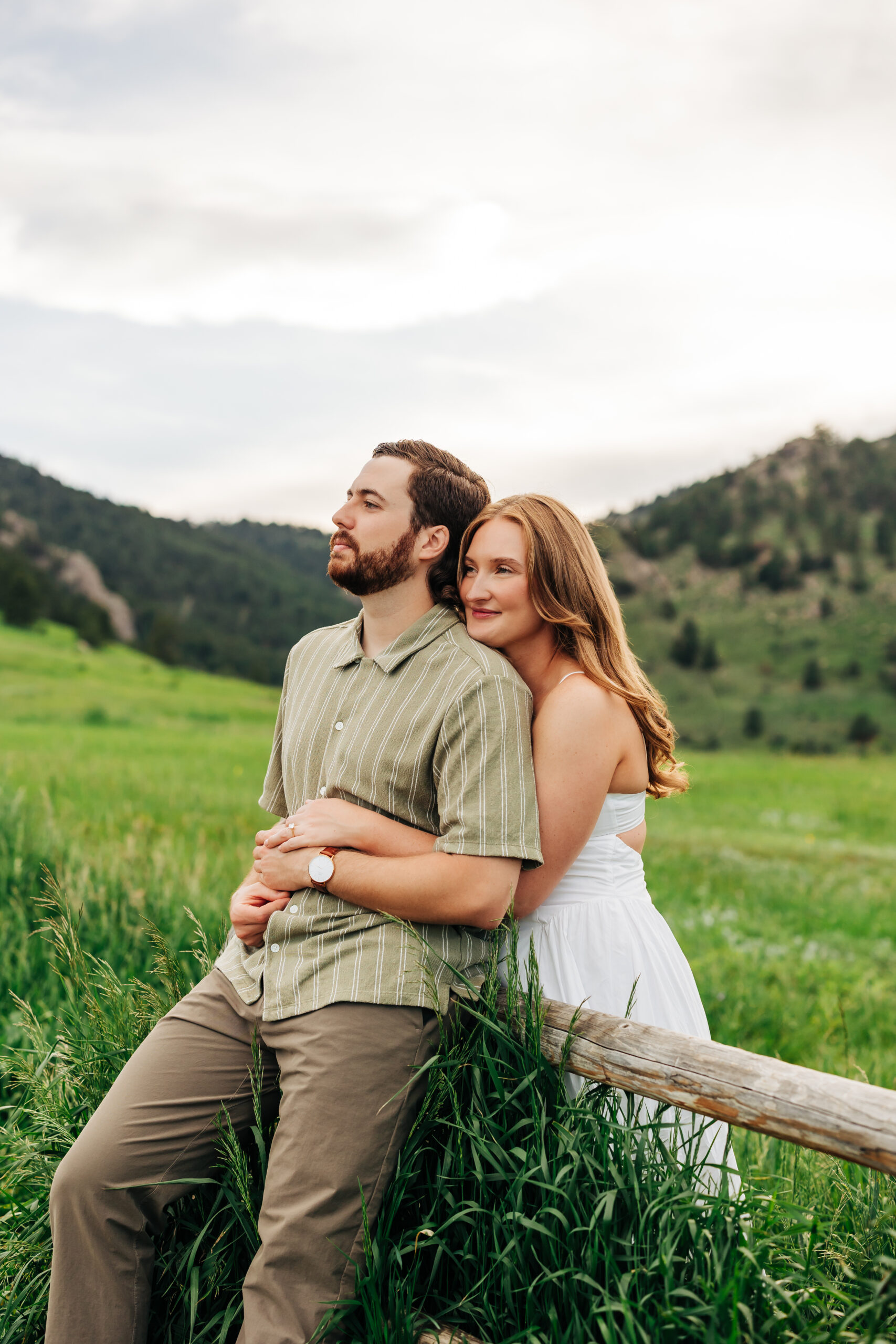 She wraps her arms around him at a rustic wooden fence, sharing a quiet moment with Boulder’s green hills as the backdrop—soft, storytelling Boulder engagement photo.