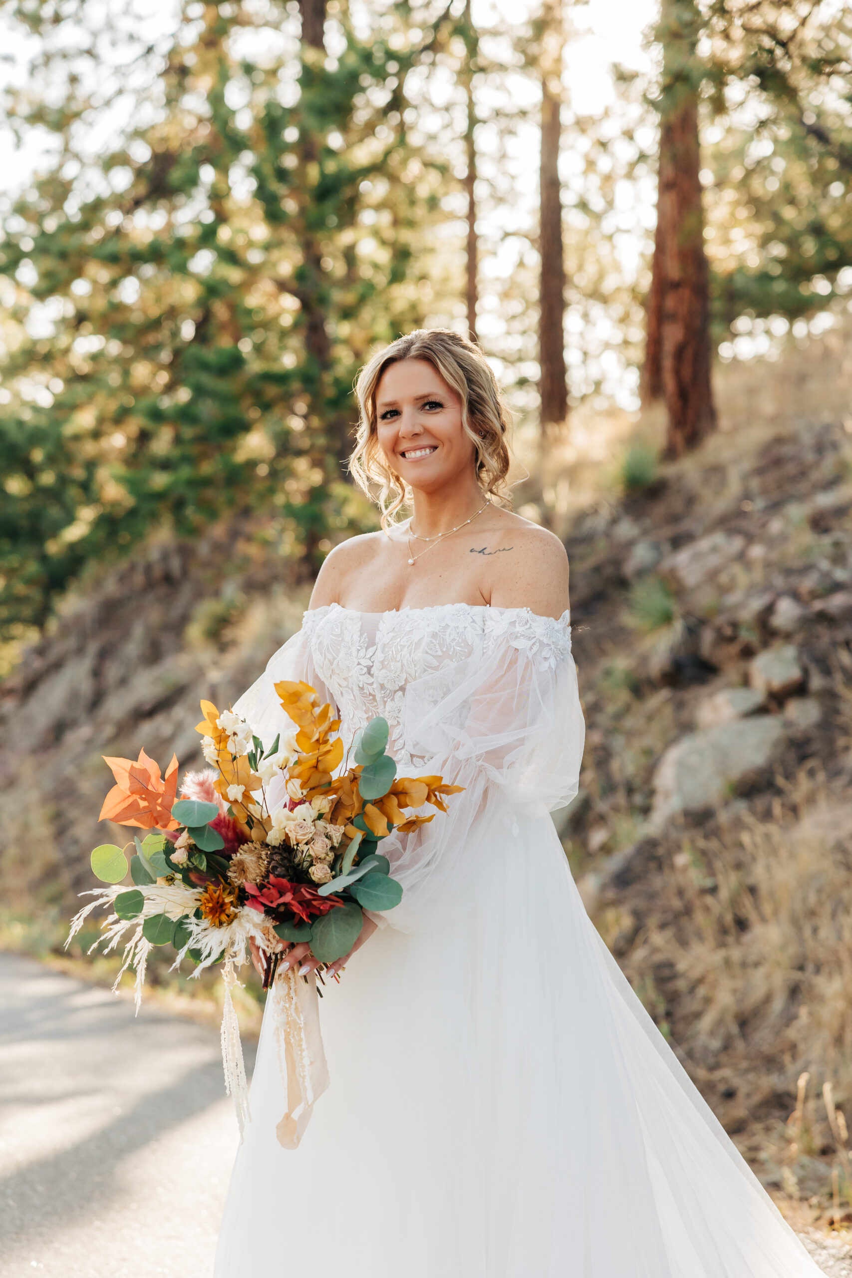 Bride glows with joy, holding a vibrant bouquet of fall florals captured by Boulder wedding photographers in a scenic woodland setting.
