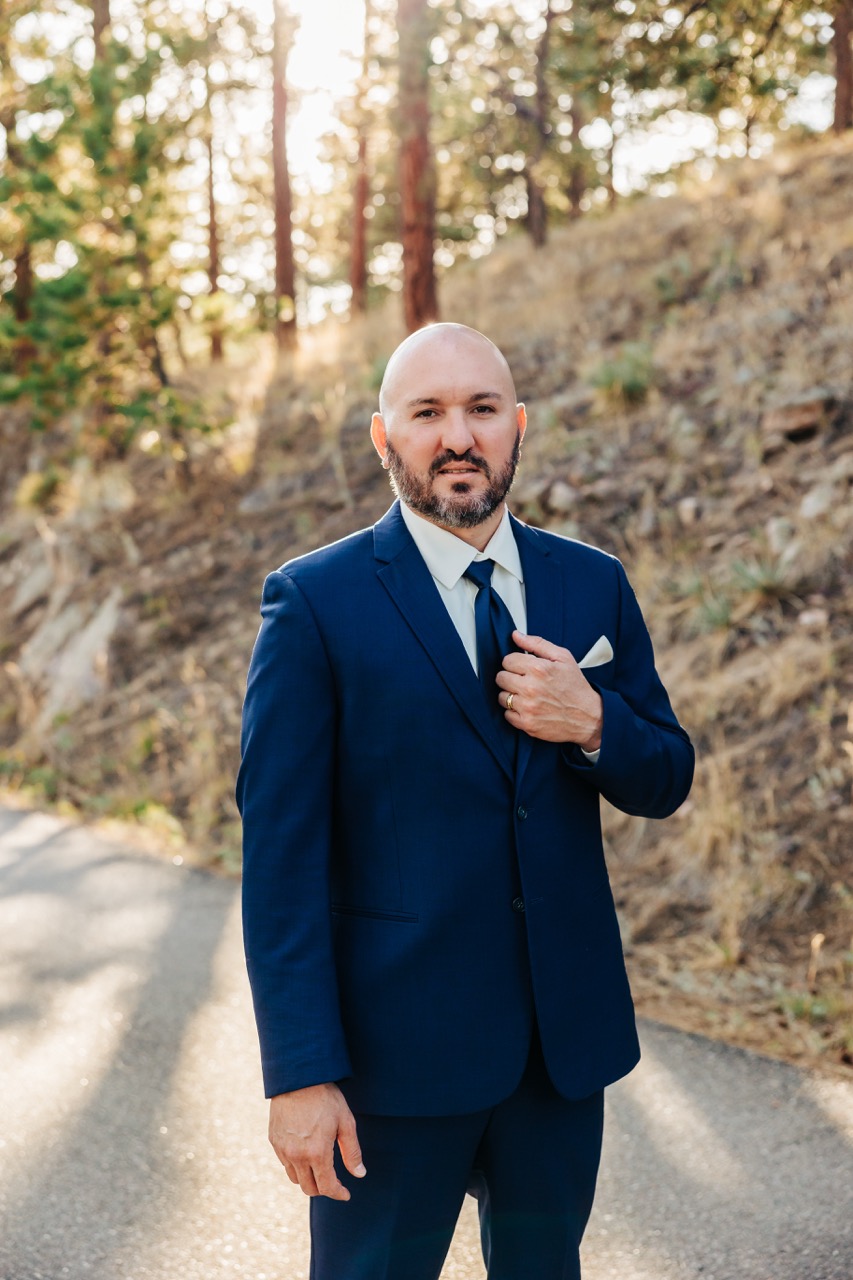 Groom in navy suit poses confidently on a forested path, sunlight streaming through the trees behind him—an elegant portrait by Boulder wedding photographers.